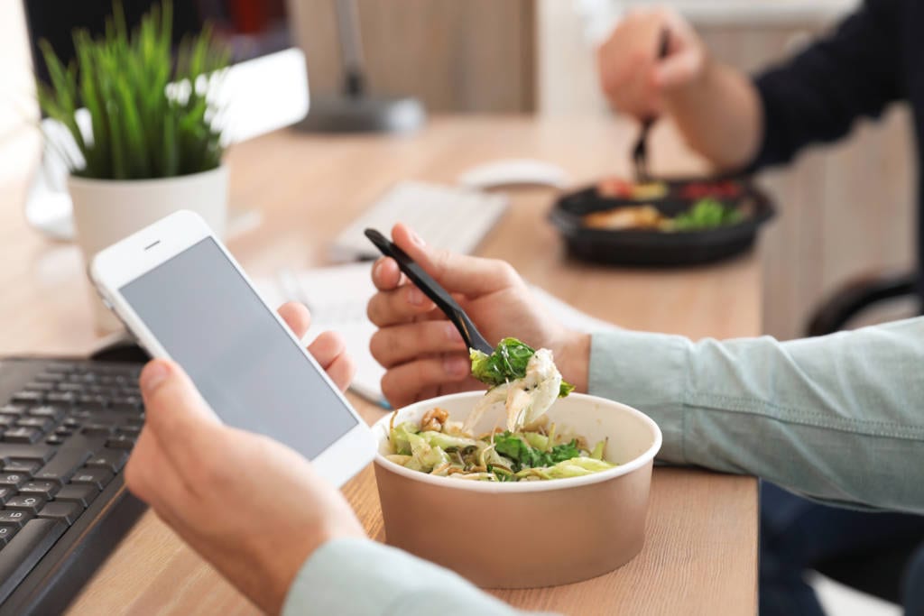 Office employee with smartphone having lunch at workplace, closeup. Food delivery